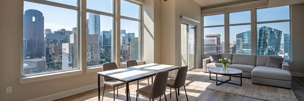 Modern apartment with large windows offering a panoramic view of a city skyline, featuring a dining table with four chairs and a living area with a gray sofa, coffee table, and a vase with flowers.