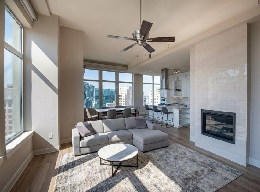 Modern living room with large windows, a gray sectional sofa, round coffee table, area rug, ceiling fan, fireplace, open kitchen, barstools, and city views through the windows. Natural light fills the space.