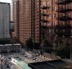 A modern outdoor pool area with lounge chairs and cabanas sits between tall brick apartment buildings in an urban setting, bathed in soft, late afternoon light.