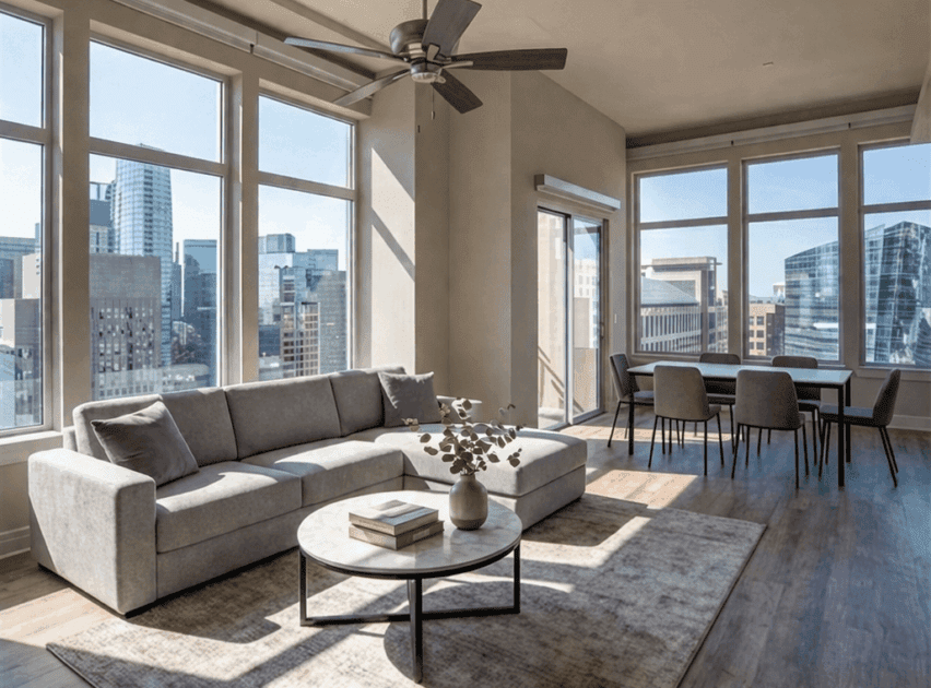A modern, bright living and dining area with large windows, a gray sofa, a round coffee table, a dining table with four chairs, and city buildings visible outside. Light wood floors and neutral decor create an airy feel.