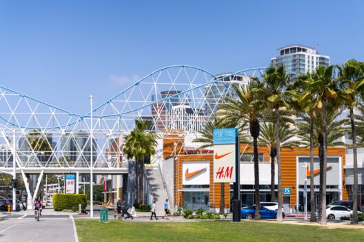 Outdoor shopping center with Nike and H&M stores, palm trees, people walking and biking, a white pedestrian bridge, and tall buildings under a clear blue sky.