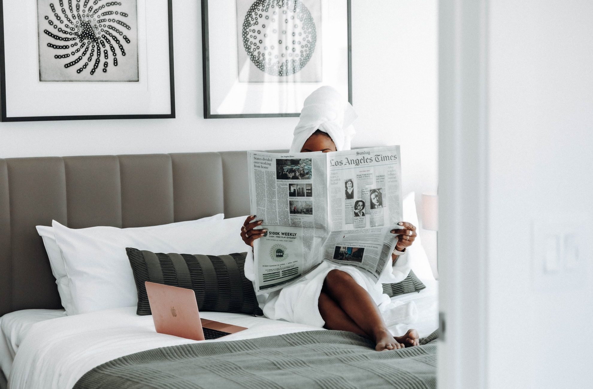 A person in a bathrobe and towel sits on a bed reading the Los Angeles Times newspaper. A closed laptop is beside them, and black-and-white art hangs on the wall above the bed.