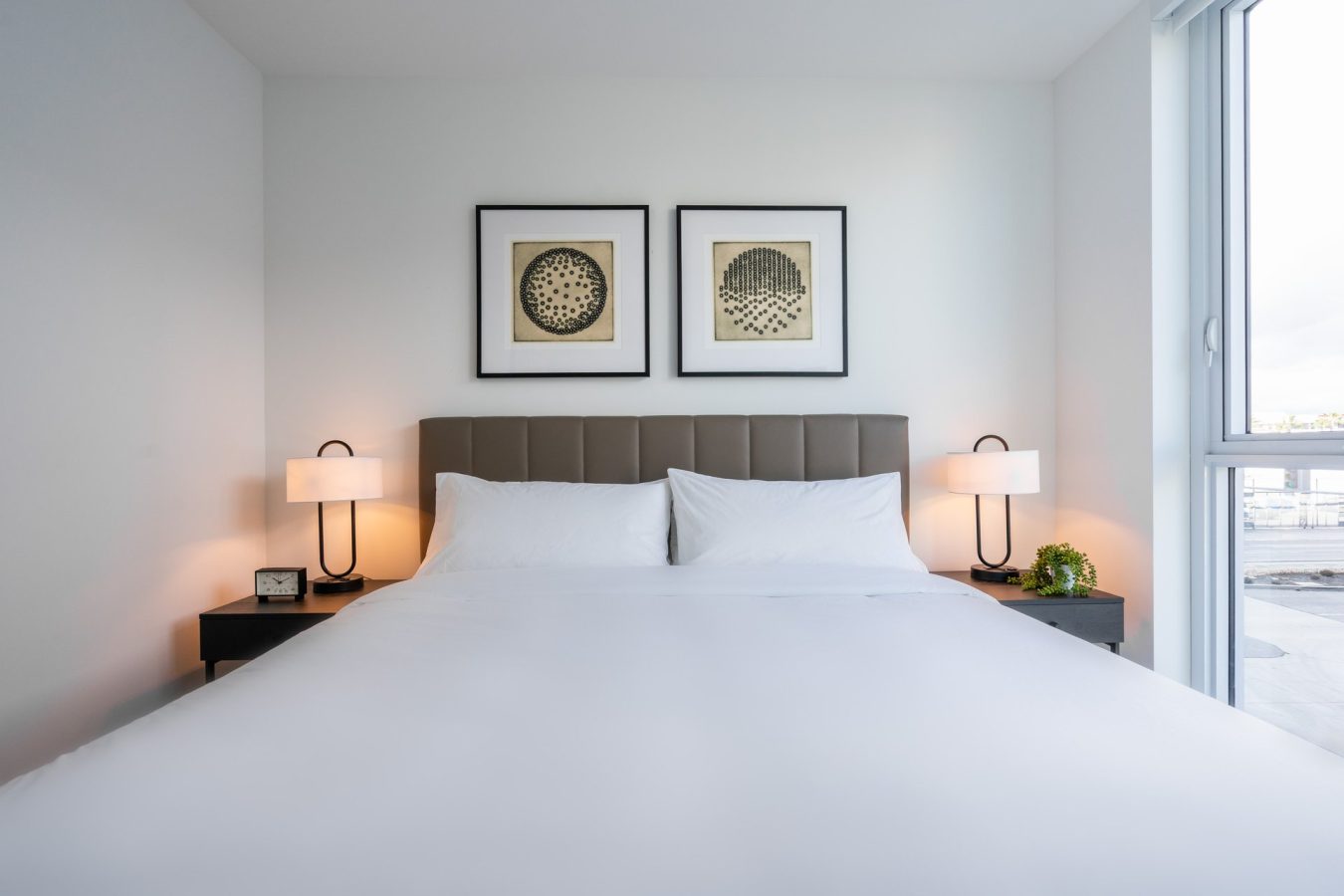 A neatly made bed with white linens, two bedside tables with lamps, a clock, and a small plant. Above the padded headboard are two framed geometric art prints. A large window lets in natural light.