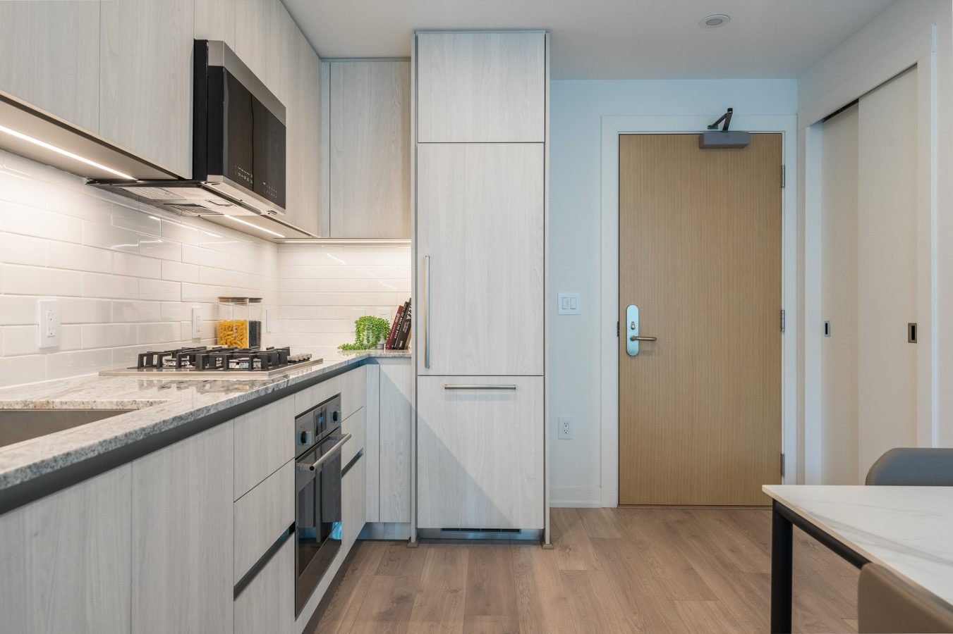 Modern kitchen with light wood cabinets, built-in appliances, a gas stove, white subway tile backsplash, and a marble countertop. A closed wooden door and a small dining table are visible on the right.