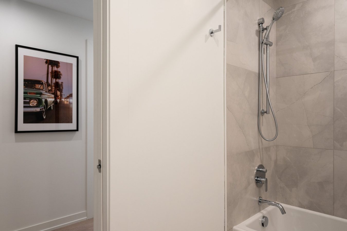 Modern bathroom with a beige tiled shower and bathtub; a handheld showerhead is mounted on the wall. In the hallway, a framed photo of a classic green car is visible on a white wall.
