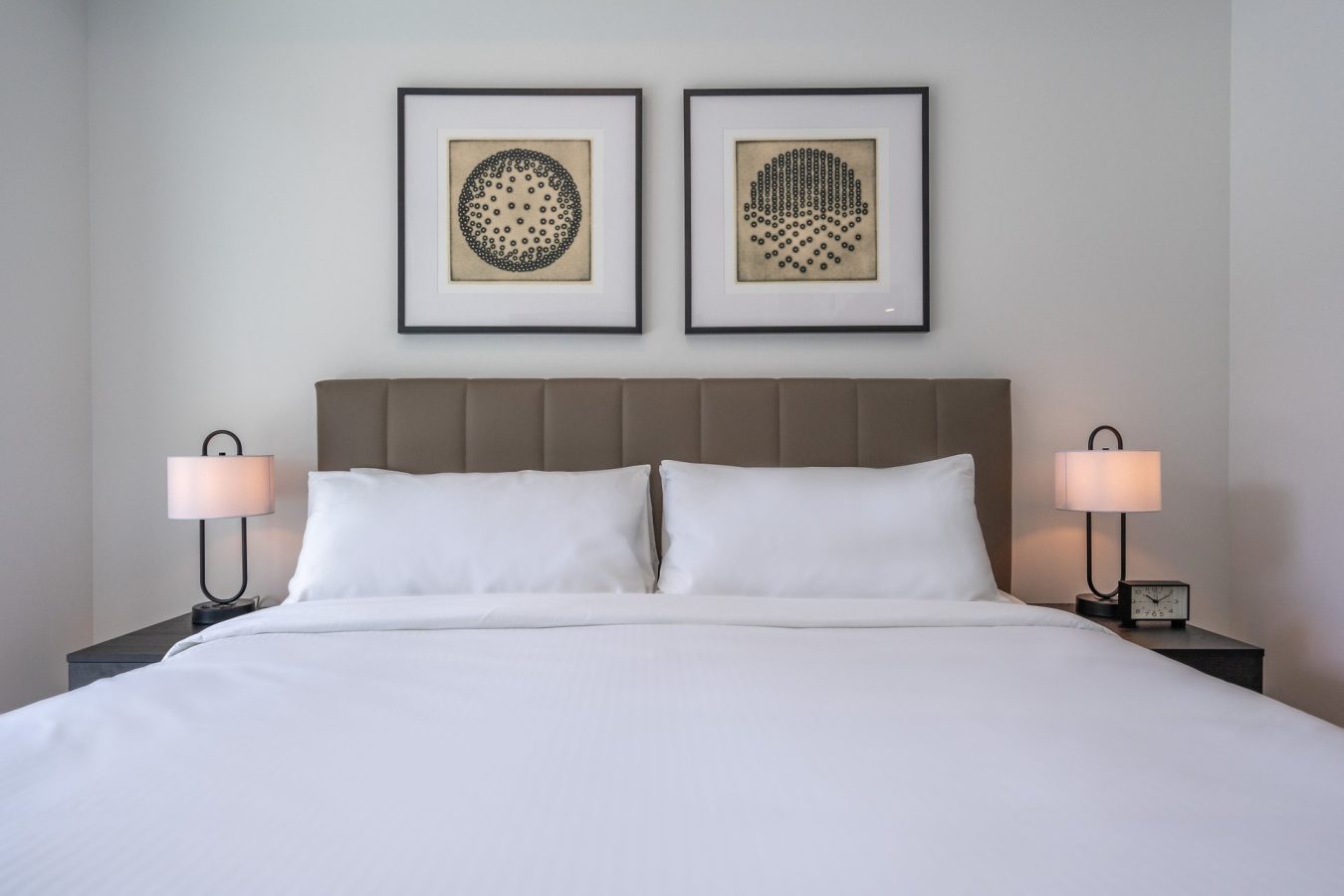 A neatly made bed with white linens, two pillows, a padded headboard, two bedside tables with modern lamps, a small clock, and two framed abstract artworks on the wall above the headboard.