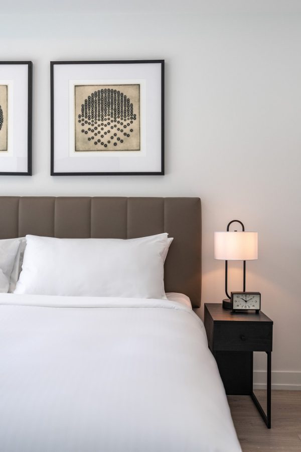 A neatly made bed with white linens is beside a black nightstand holding a clock and a lamp. Two framed abstract artworks hang above the upholstered headboard against a white wall.
