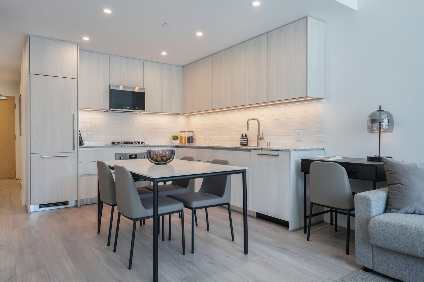 Modern kitchen with light wood cabinets, integrated appliances, a black dining table with four gray chairs, a countertop with a bowl of fruit, and a small desk area next to a gray sofa. Bright, minimalist design.