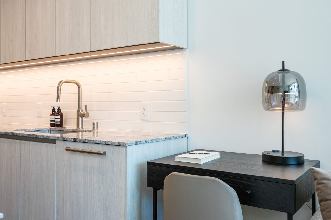 A modern kitchen area with light wood cabinets, a marble countertop, a sink, soap dispensers, and a white tile backsplash. Next to it, there’s a dark desk with a book, chair, and a glass lamp.