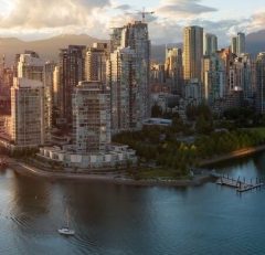 City skyline with tall modern buildings along the waterfront, a small sailboat in the water, green parkland, and distant mountains under a partly cloudy sky at sunset.
