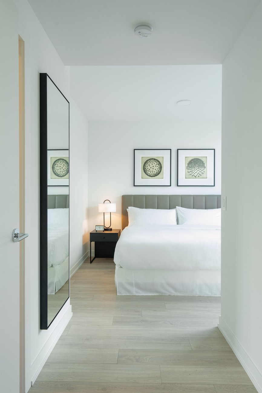 A modern, minimal bedroom with a large bed covered in white linens, a black nightstand with a lamp, two framed geometric prints on the wall, light wood flooring, and a tall mirror on the left wall.