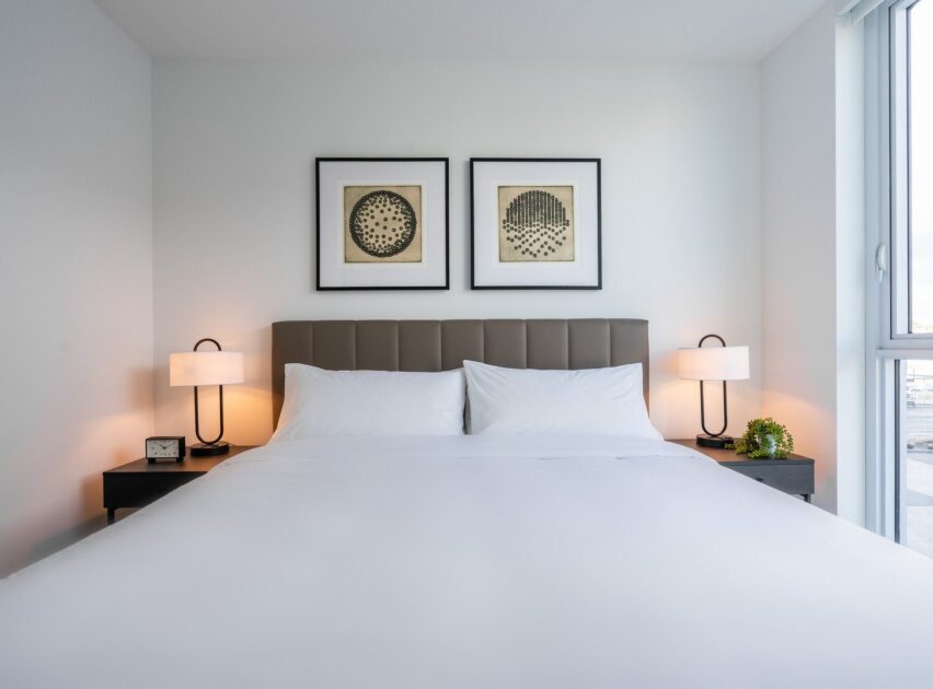 A modern, minimal bedroom with a neatly made bed, two white pillows, bedside tables with lamps, a clock, and a plant. Above the bed are two framed abstract artworks. Natural light comes through a large window on the right.