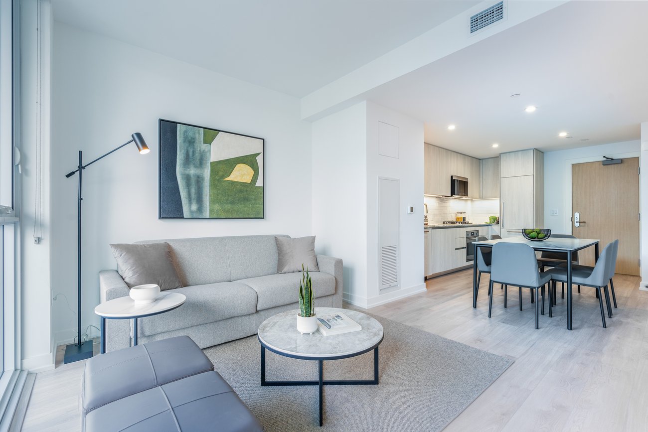 Modern open-plan living room and kitchen with light wood flooring, gray sofa, round coffee table, dining area, and contemporary wall art. Space is bright with natural light coming through large windows.