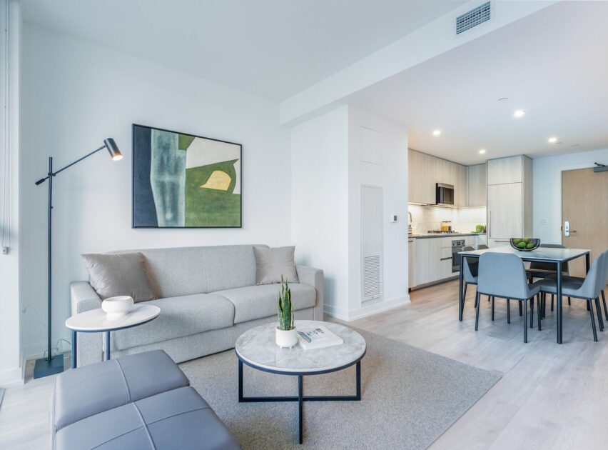 Modern, bright living room with a gray sofa, round coffee table, floor lamp, and abstract art on the wall. The space opens to a kitchen and dining area with light wood cabinets and a dining table with four chairs.
