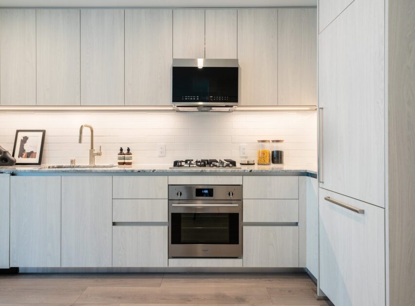 Modern kitchen with light wood cabinets, stainless steel oven and stove, a black range hood, and white subway tile backsplash. Countertop holds soap dispensers, a framed picture, and a jar with dry pasta.