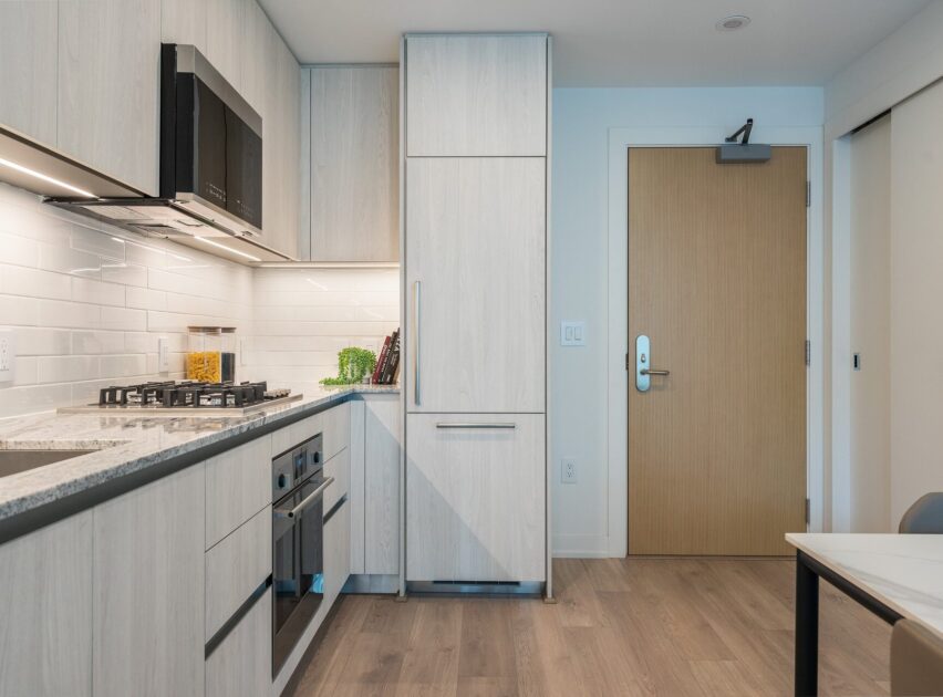 Modern kitchen with light wood cabinets, built-in oven and microwave, gas stovetop, backsplash of white subway tiles, and a closed wooden door. Countertops hold a few kitchen items. Floors are light wood.