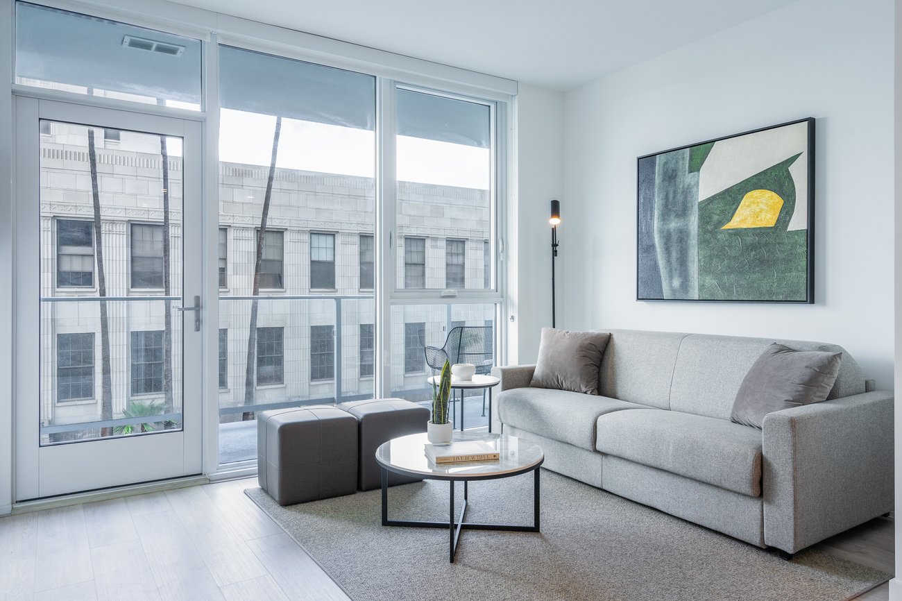 Modern living room with large windows, a light gray sofa, abstract wall art, a round coffee table with a plant, two ottomans, a floor lamp, and city building views outside.