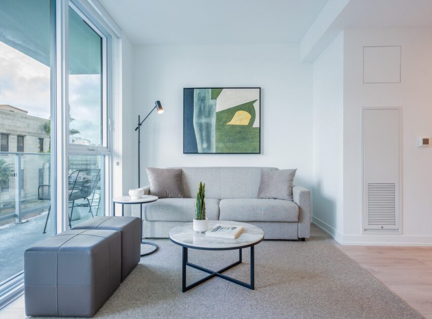 A modern living room with a light gray sofa, two round tables, a black floor lamp, a small plant, and abstract art on the wall. Large glass doors open to a balcony with outdoor seating. The room is bright and minimalist.