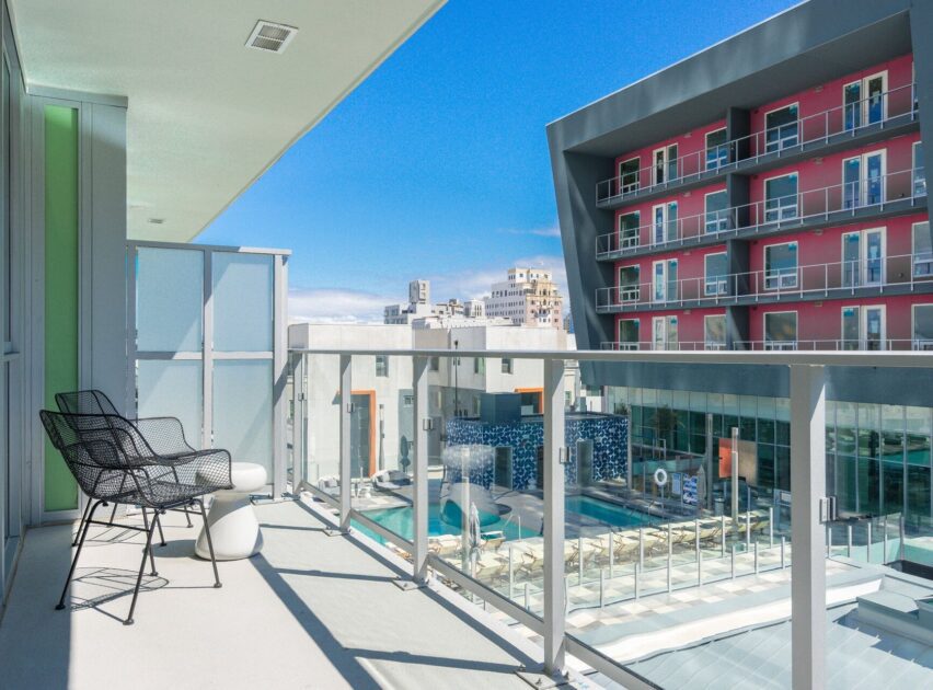 A modern balcony with two black chairs and a small table overlooks a pool area and a red and gray apartment building on a sunny day. Glass railing offers an unobstructed city view.
