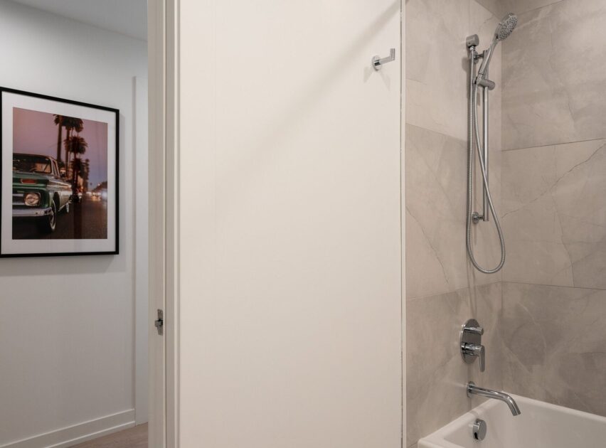 A modern bathroom with beige marble tiles, a wall-mounted showerhead, and a bathtub. An open door reveals a hallway with a framed photograph of a vintage green car and palm trees.