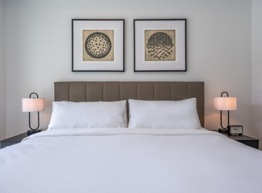 A neatly made bed with white linens, two pillows, and a padded headboard. On each side are small bedside tables with modern lamps. Two framed abstract artworks hang on the wall above the headboard.