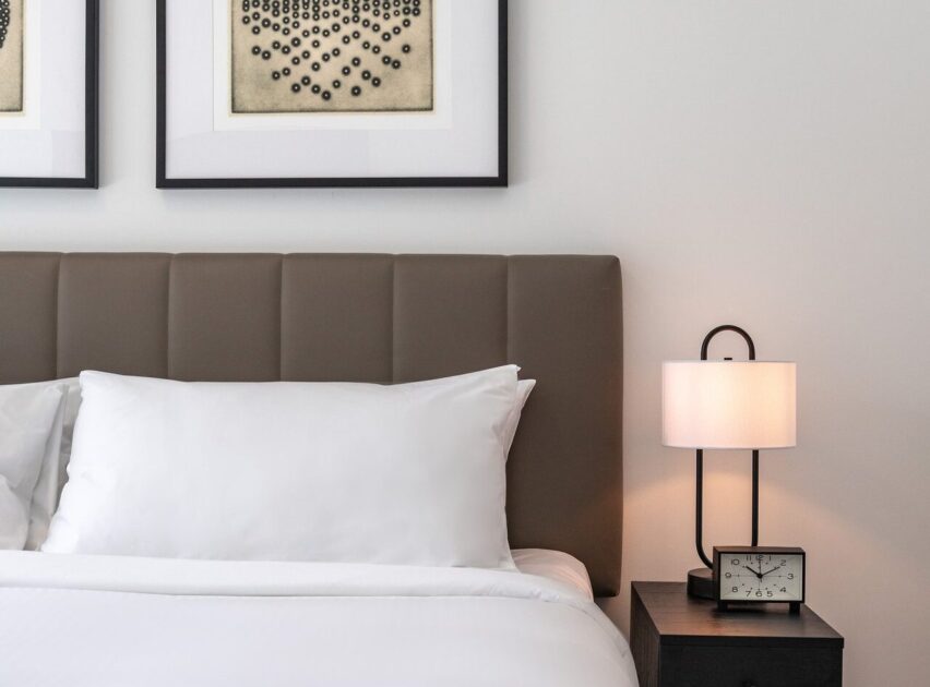 A neatly made bed with white linens and a padded headboard next to a nightstand holding a modern lamp and a small clock, against a white wall with framed abstract artwork above.