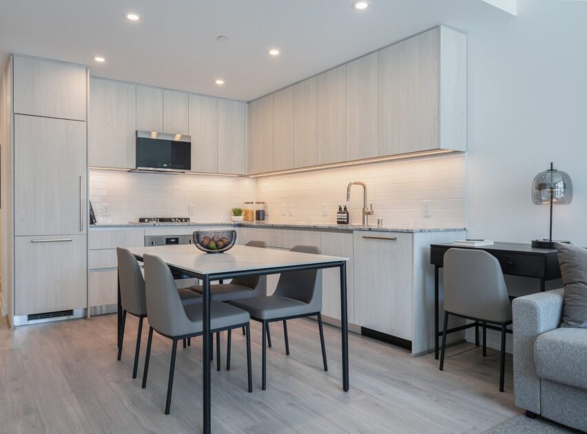 Modern kitchen with light wood cabinets, built-in appliances, a dining table with four gray chairs, and a small black desk with a chair in the corner. The space has light flooring and under-cabinet lighting.