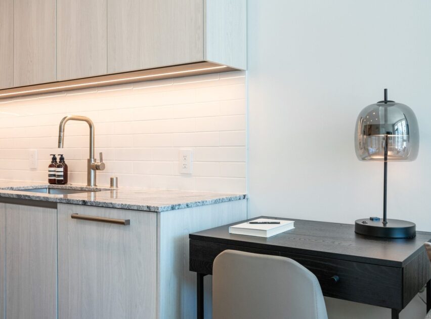 A modern kitchenette with light wood cabinets, a marble countertop, a stainless steel faucet, white tile backsplash, and two soap bottles next to a dark desk with a lamp, chair, and a closed book.