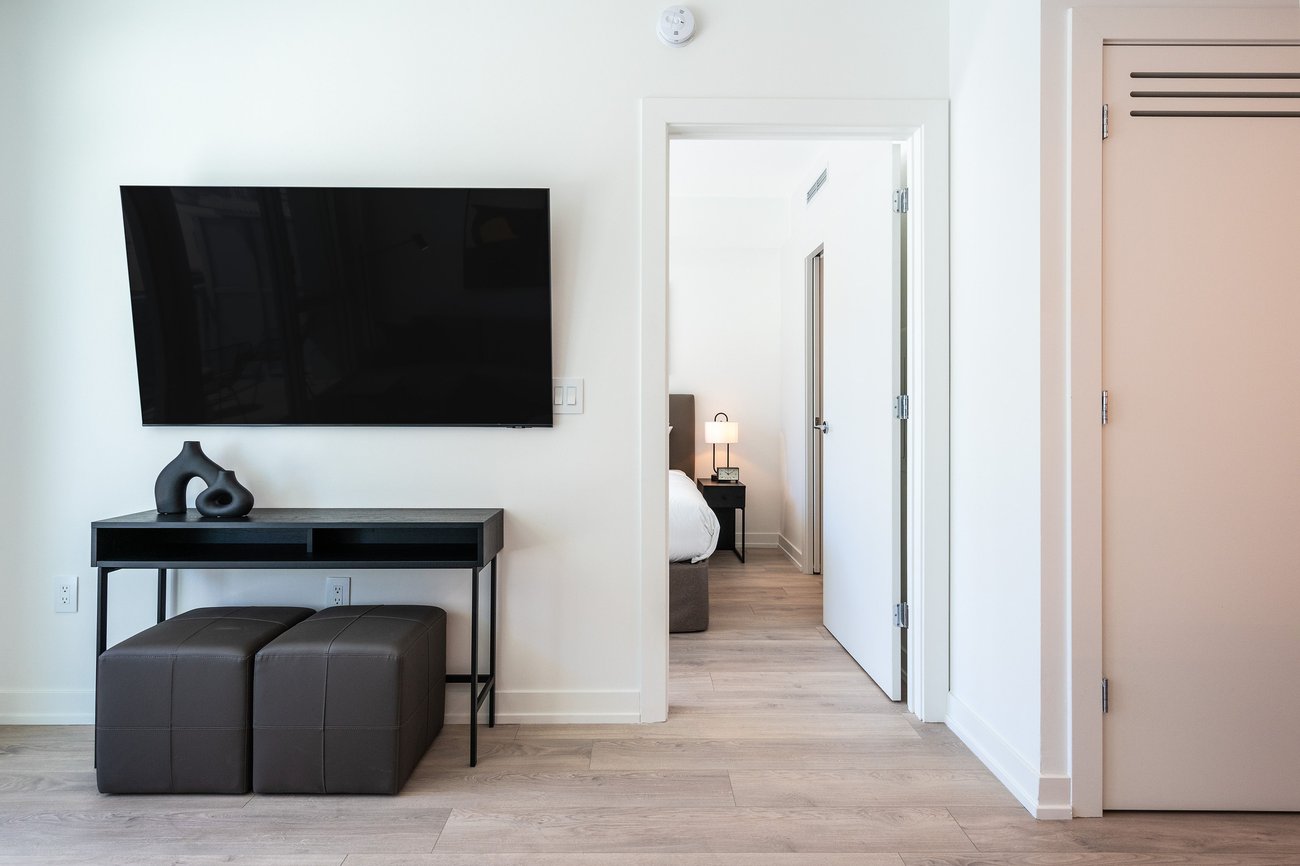 Minimalist living space with a wall-mounted flat-screen TV above a black console table, two brown ottomans, light wood flooring, an open door to a bedroom, and a closet door on the right.