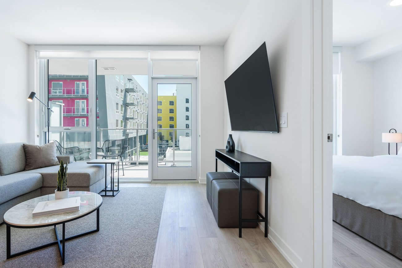 A modern, bright apartment living room with a gray sofa, round coffee table, large wall-mounted TV, and sliding glass doors leading to a balcony. A bedroom with a bed and lamp is visible through an open doorway.