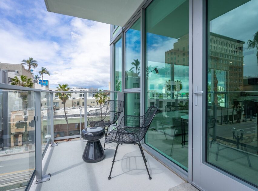 A modern balcony with two black mesh chairs and a small round table overlooks a city street lined with palm trees and buildings, with reflections visible in the glass windows and doors.