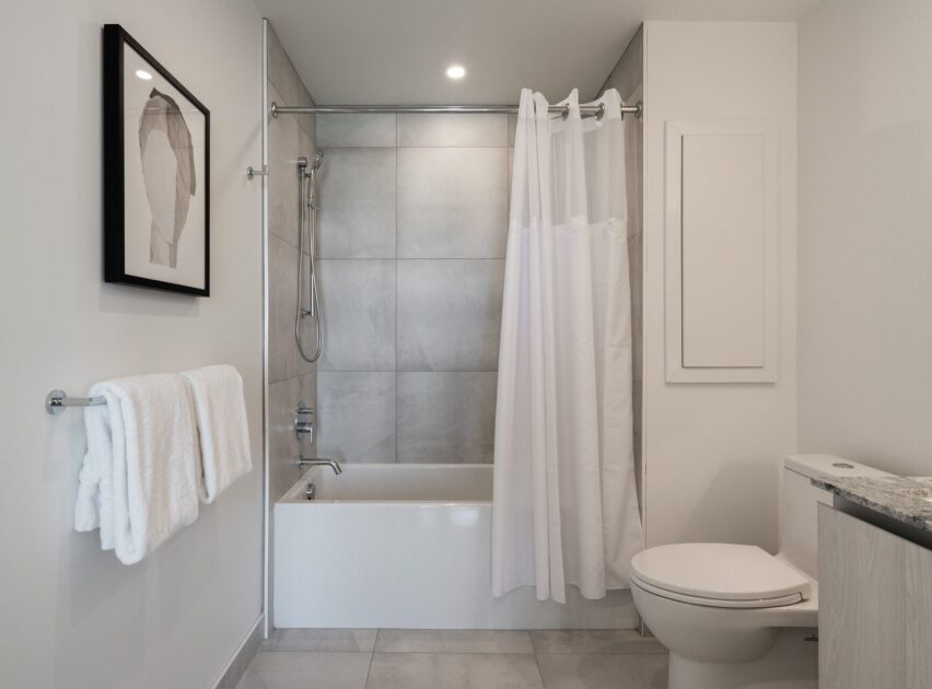 Modern bathroom with a white bathtub and shower combo, gray tile walls, a white shower curtain, toilet, towel rack with white towels, and abstract art on the wall. Neutral color scheme and clean design.