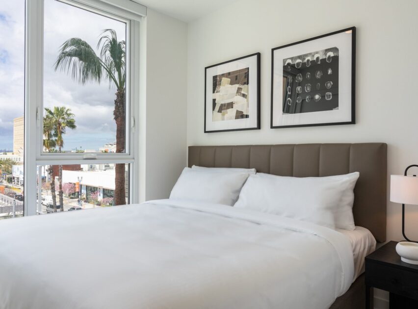 A modern bedroom with a large bed covered in white linens, two abstract framed artworks above the headboard, a nightstand with a lamp, and a window showing palm trees and city buildings outside.