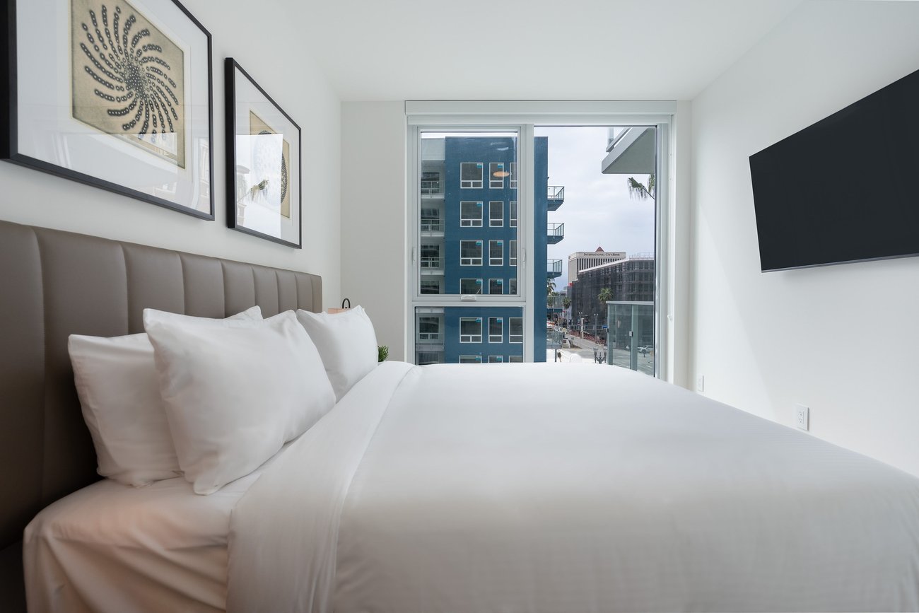 A modern, bright bedroom with a neatly made bed, white linens, framed artwork on the wall, a large window with city views, and a wall-mounted TV.