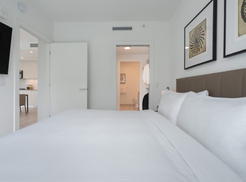 A modern bedroom with a neatly made bed, white linens, two framed artworks above the headboard, and an open door revealing a bathroom and part of a kitchen area in the background.
