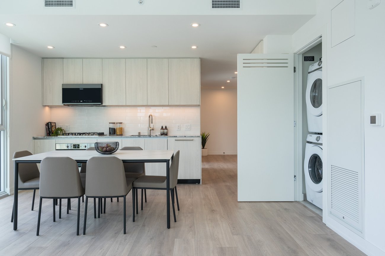 Modern kitchen and dining area with light wood cabinets, a black dining table with six brown chairs, and a stackable washer and dryer in an open closet. Bright, minimalist design with light wood flooring.