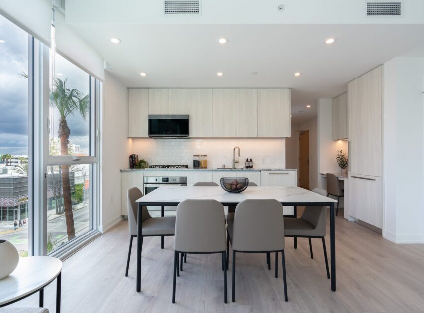 Modern kitchen with light wood cabinets, a white tile backsplash, built-in appliances, and a white dining table with six beige chairs. Large windows let in natural light, and a palm tree is visible outside.