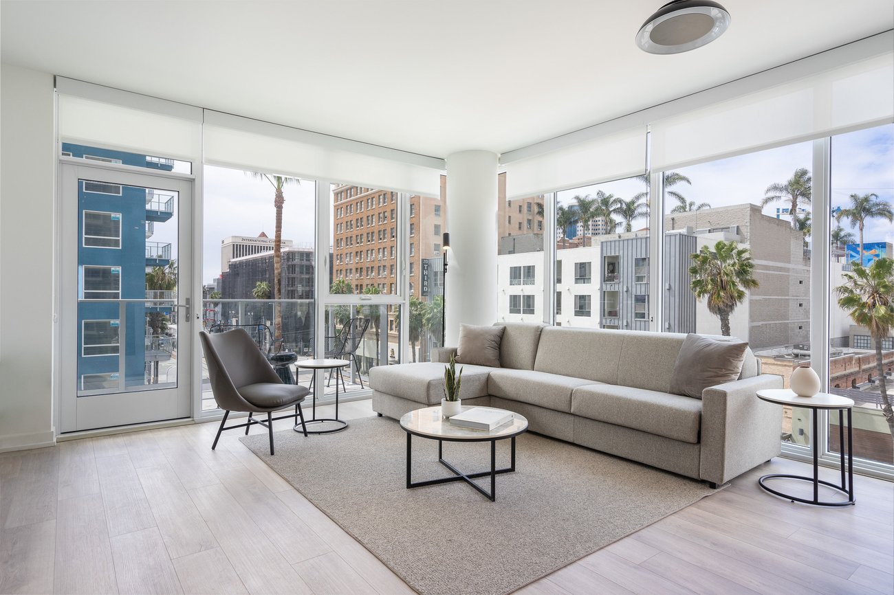 A modern, bright living room with large windows, a gray sofa, a black chair, two round coffee tables, light wood floors, and city buildings with palm trees visible outside.