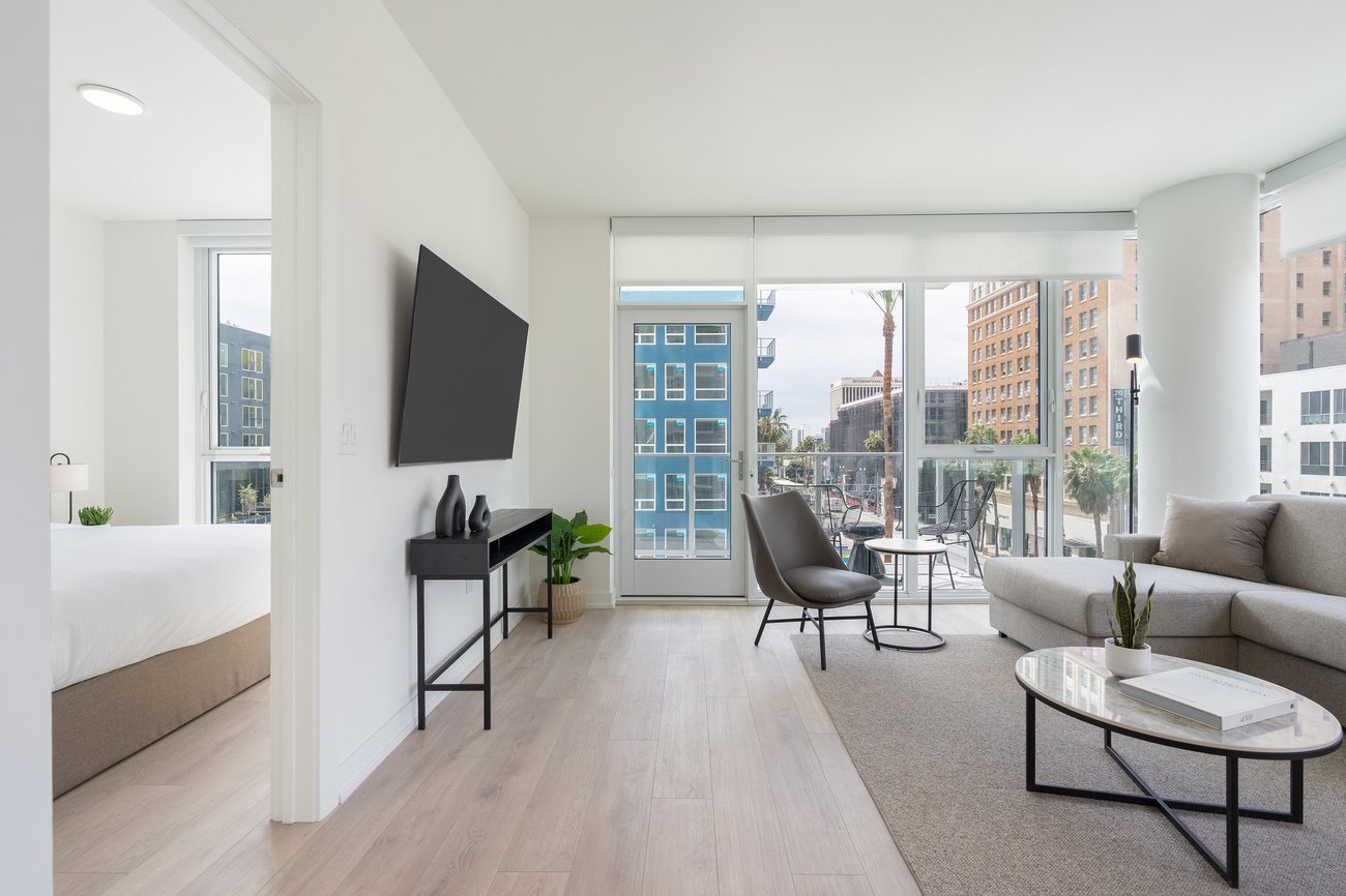 Modern, bright apartment with a living room featuring a sofa, armchair, coffee table, TV, and balcony access. A bedroom with a neatly made bed is visible through an open doorway. Large windows let in natural light.