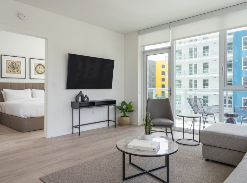 A modern, bright apartment living room with a wall-mounted TV, round coffee table, gray chair, and a small console table. Large windows reveal a balcony and neighboring buildings. A bedroom is visible through an open doorway.
