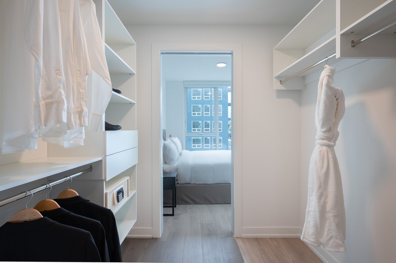 A modern walk-in closet with white shelves and hanging clothes leads to a bright bedroom with a neatly made bed and large window overlooking city buildings. A white robe hangs on the right.