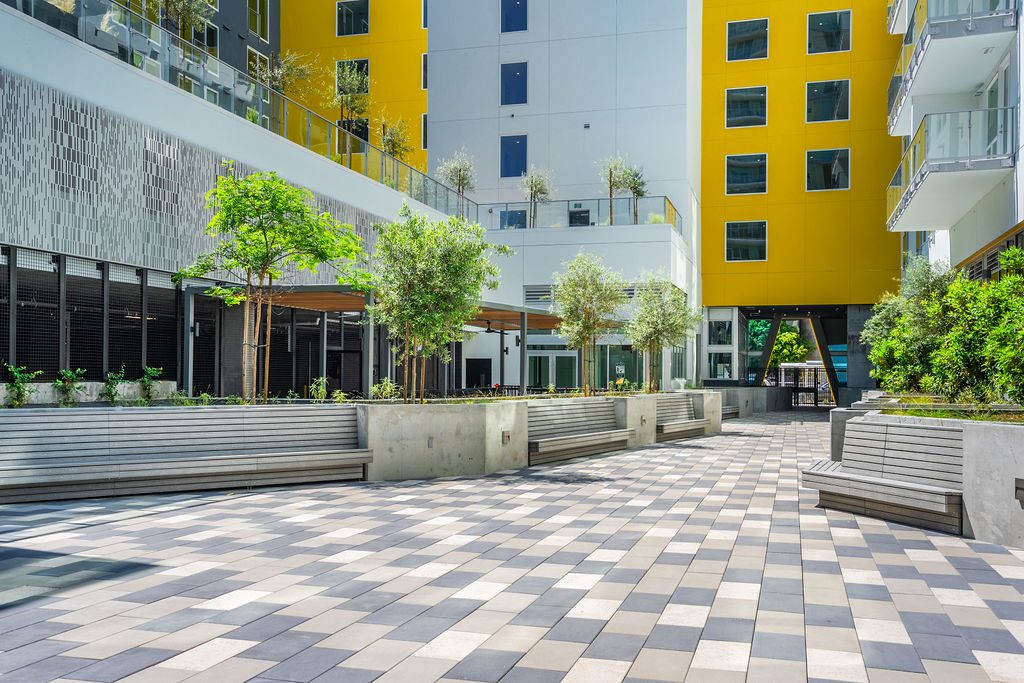 A modern urban courtyard with patterned tile flooring, benches, young trees, and surrounding yellow and white apartment buildings with balconies and glass railings.