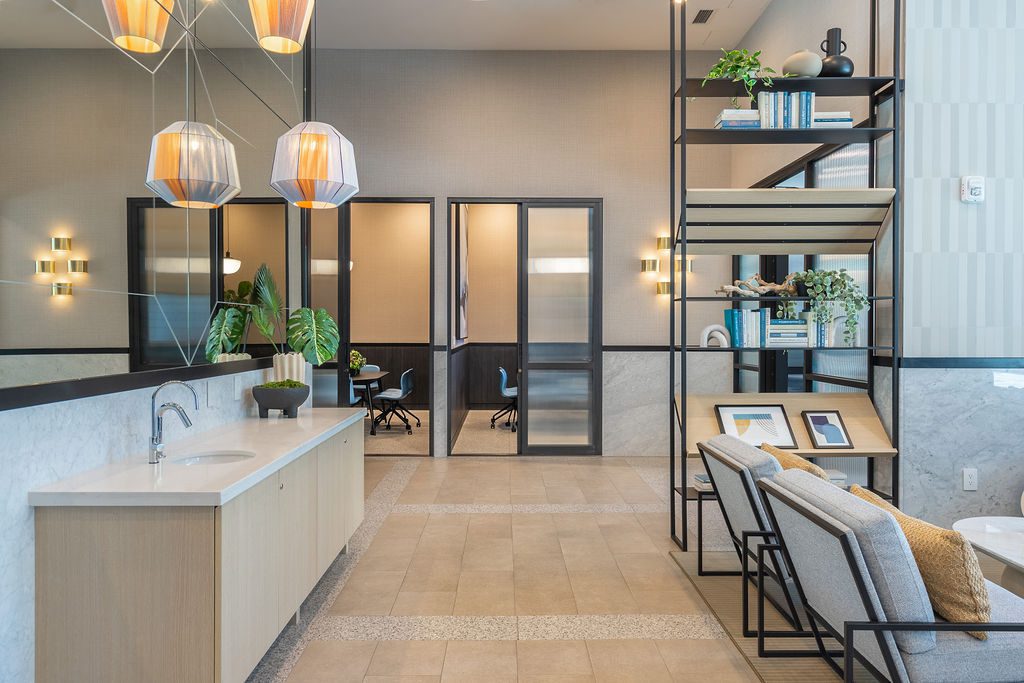 Modern office lounge with light wood cabinets, a sink, two pendant lights, a tall shelving unit with books and decor, and two armchairs. Glass doors reveal a meeting room in the background.