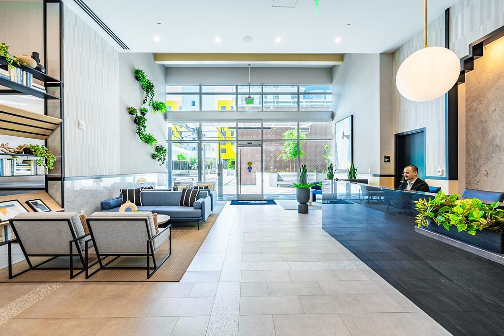 Modern lobby with stylish seating, plants, large windows bringing in natural light, and a receptionist at a sleek black front desk. Shelves with decor and greenery enhance the contemporary space.