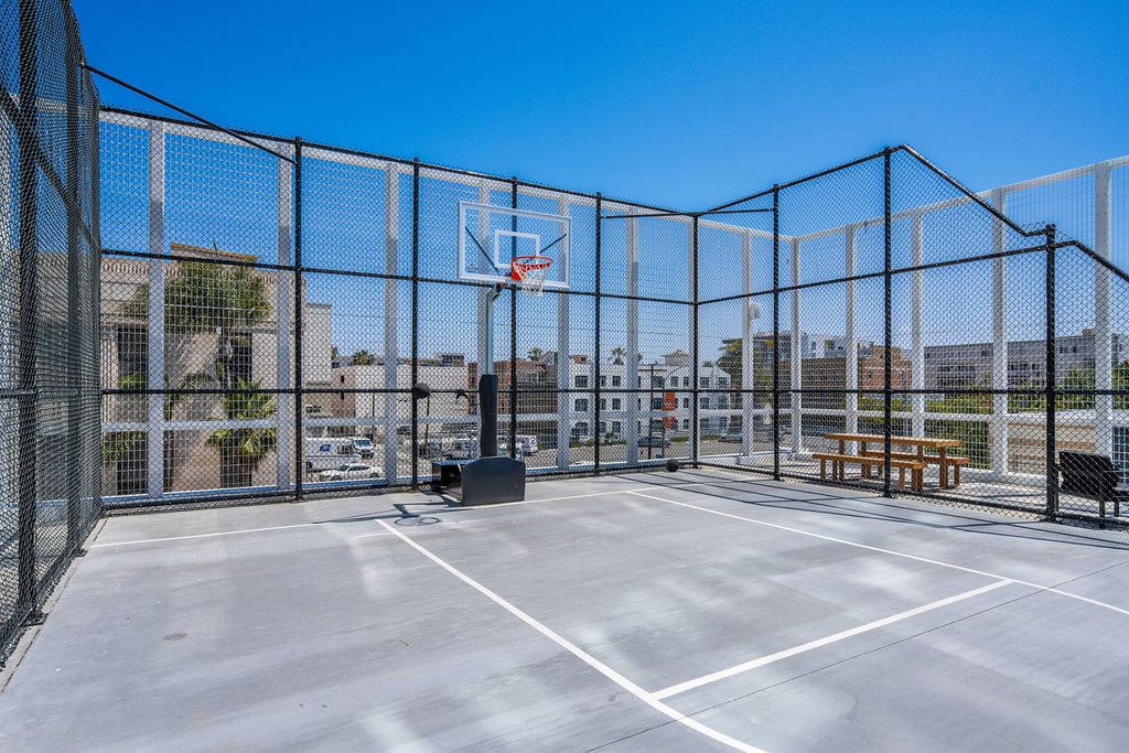 An outdoor rooftop basketball court enclosed with wire fencing, featuring a hoop, court markings, and a wooden picnic table on the side. Buildings and blue sky are visible in the background.
