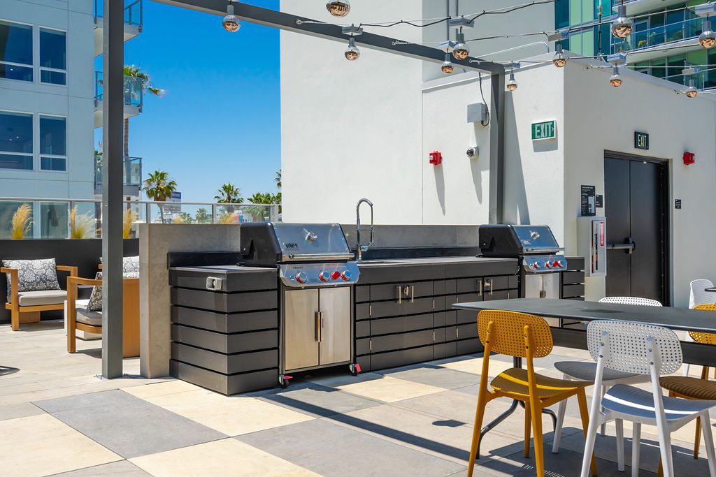 Modern outdoor kitchen with two stainless steel grills, a sink, and black cabinetry. There are yellow and white chairs around a dark table, string lights overhead, and palm trees visible in the background.