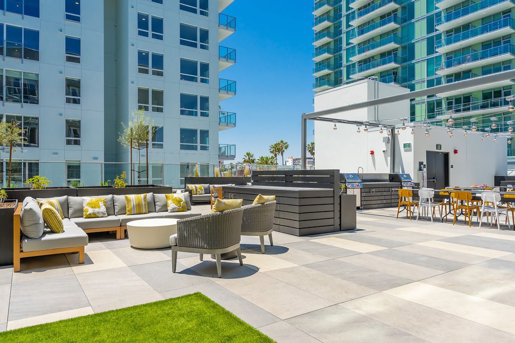 Modern outdoor patio with cushioned seating, round tables, string lights, and dining area between tall glass apartment buildings under a clear blue sky. Green artificial grass is in the foreground.