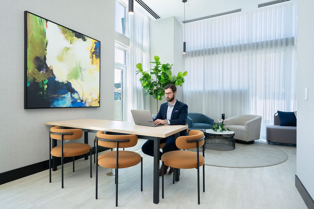 A man in business attire works on a laptop at a modern table with four orange chairs in a bright, stylish office space. Large abstract art, a potted plant, and lounge seating are visible in the background.