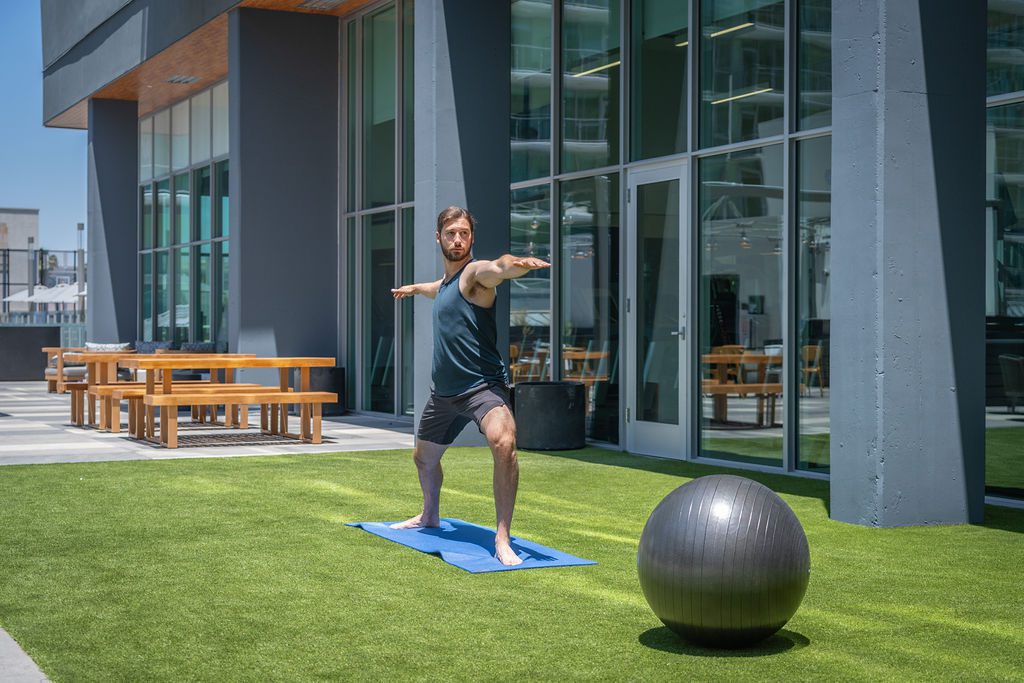 A man practices yoga in Warrior II pose on a blue mat outdoors, near a modern building with large windows. A gray exercise ball is on the grass nearby, and wooden picnic tables are in the background.