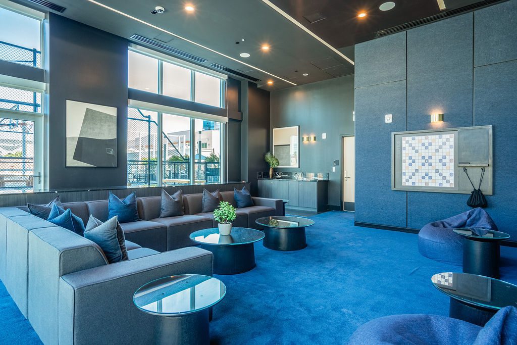 A modern lounge with blue carpet, gray sectional sofas, round glass tables, blue beanbags, wall art, a board game wall installation, and large windows letting in natural light.